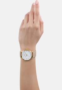 Pols met een beige leren bandhorloge met een witte ronde wijzerplaat, gouden kast en minimalistische gouden uurmarkeringen en wijzers.