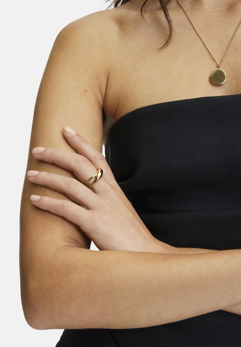 Gouden gedraaide ring om een hand, gedragen met een strapless zwart kledingstuk. Huidtextuur zichtbaar op de arm, met een eenvoudige gouden hangerketting.