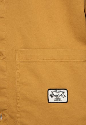 Beige canvasjacka med en rektangulär patch med en logotyp som säger "DC Shoe Company Worldwide Since 1994" på bröstfickan.