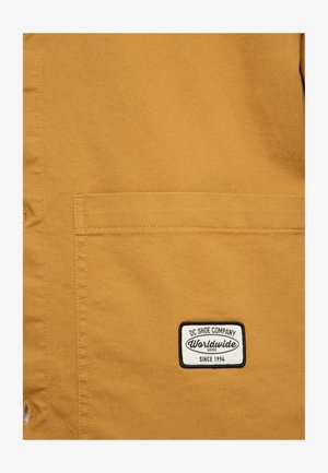 Beżowa kurtka z płótna z prostokątną naszywką z logo "DC Shoe Company Worldwide Since 1994" na kieszeni piersiowej.