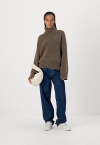 Maglione marrone, di grandi dimensioni, in maglia con collo alto; pantaloni in denim larghi; borsa color crema; sneaker bianche; tessuto strutturato e vestibilità rilassata.