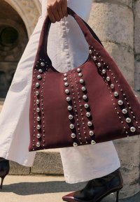 Bordo platnena torba s metalnim srebrnim zakovicama u vertikalnim linijama i okruglim otvorom, u kombinaciji s bijelim hlačama i crnim potpeticama.