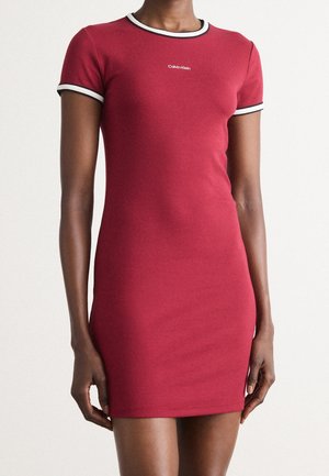 Woman wearing fitted short-sleeve red dress with white and black trim on sleeves and collar, small white Calvin Klein logo on chest.
