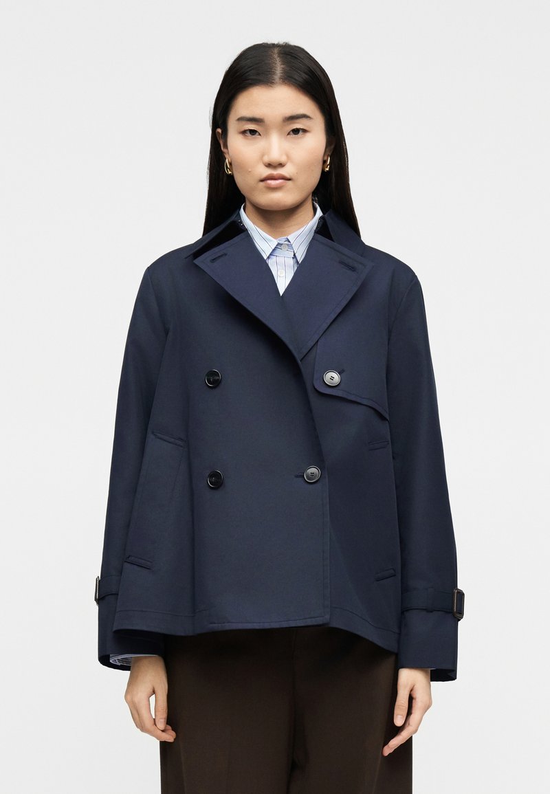 Γυναίκα με ίσια σκούρα μαλλιά, φορώντας χρυσούς  κρίκους σκουλαρίκια, ναυτικό διπλόμπρατσο σακάκι, μπλε ριγέ πουκάμισο και σκούρο παντελόνι, στραμμένη προς τα εμπρός.