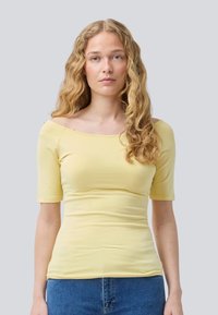 Gele top met korte mouwen en een off-shoulder halslijn, strakke pasvorm en zachte stoffen textuur, gecombineerd met blauwe denim jeans.