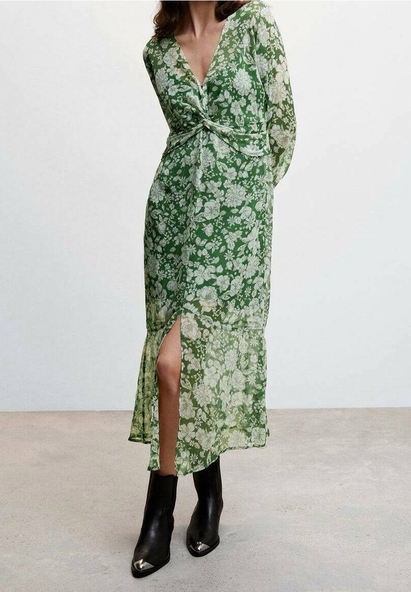 Femme portant une robe longue à manches longues verte à motif floral avec une fente avant et un détail noué, associée à des bottines noires à bouts argentés.