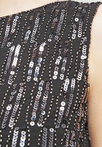 Černá tkanina s kaskádovými řadami metalických flitrů a korálků, vytvářejícími texturovaný, třpytivý vzor. Detailní pohled na oblast ramene.