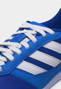 Baskets de sport bleues et blanches avec des lacets blancs, trois bandes blanches sur le côté et une étiquette bleue sur la languette avec un texte partiellement visible.