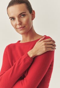 Sarkana, adīta džemperis ar apaļu kakla izgriezumu un ribotu piedurkņu galotni, ar mīkstu tekstūru. Modelis atpūš roku uz pleca.