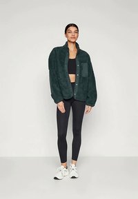 Mørkegrøn, fuzzy fleece skjortejakke med knaplukning og en brystlomme, båret over en sort crop top og sorte leggings.