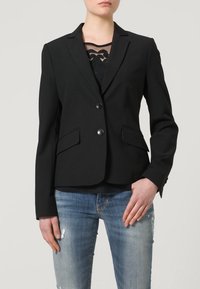Blazer noir ajusté avec deux boutons à l'avant, revers crantés et poches avant, associé à un haut en dentelle noire et un jean bleu déchiré.