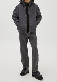 Veste à capuche grise zippée avec détail logo, chemise noire, pantalon en jean gris et chaussures noires à lacets. Tissu lisse et coupe décontractée.