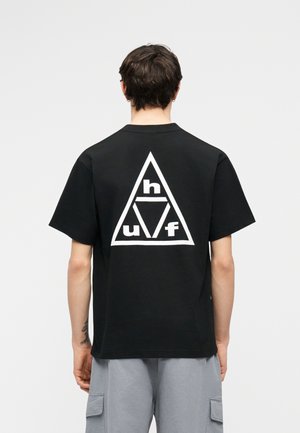 Άνδρας που φοράει μαύρο μπλουζάκι με μεγάλο λευκό τριγωνικό γραφικό και τα γράμματα «h», «u» και «f» στην πλάτη, συνδυασμένο με γκρι cargo σορτς.