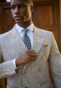 Abito beige in lino doppiopetto con leggere righe, abbinato a una cravatta blu a paisley. Un fazzoletto da taschino spunta dalla tasca del petto.