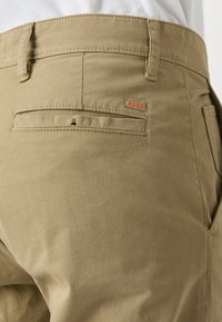 Gros plan sur la poche arrière d’un pantalon beige avec passants de ceinture et petite étiquette orange « BOSS » cousue au-dessus de la poche.