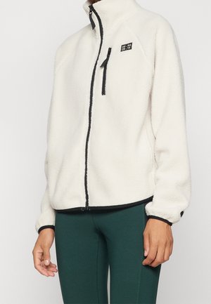 Osoba ubrana w biały polar z czarnymi detalami na zamku błyskawicznym i małą naszywką na piersi, w połączeniu z ciemnozielonymi spodniami.