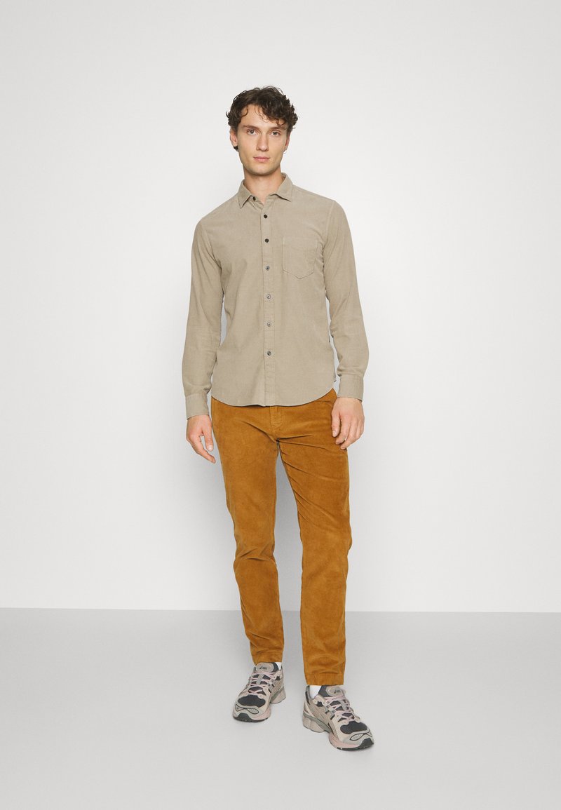 Chemise beige à boutons avec une poche avant, associée à un pantalon en velours côtelé marron. Portée avec des baskets grises et noires. Fond minimaliste.