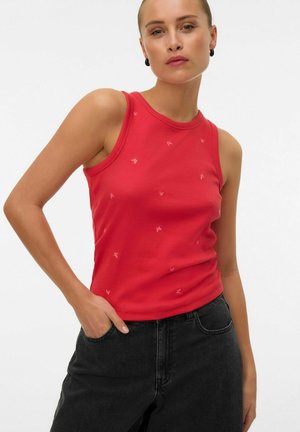 Vrouw met een rood mouwloos topje met een subtiel vlinderpatroon en zwarte spijkerbroek, poserend met één hand in de zak tegen een effen achtergrond.