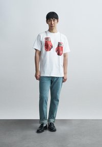 Λευκό T-shirt με γραφικό κόκκινων γαντιών πυγμαχίας. Το μοντέλο φοράει μπλε τζιν και μαύρα παπούτσια με λευκές κάλτσες. Απλός, καθημερινός σχεδιασμός.