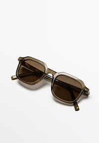 Kwadratowe okulary przeciwsłoneczne z przezroczystego szaro-brązowego plastiku, z ciemnymi przyciemnianymi szkłami i subtelnym teksturowanym wzorem na zausznikach.