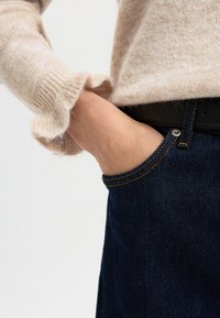 Maglione beige a maglia con maniche volantee, abbinato a jeans in denim scuri con cuciture gialle e una cintura in pelle nera. Dettaglio della mano in tasca.