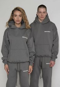 Conjunto de sudadera con capucha y pantalones deportivos grises, con bolsillos tipo canguro y cordón ajustable. Logo blanco con texto "MULTIPLY" en el pecho y el muslo. Tela suave.