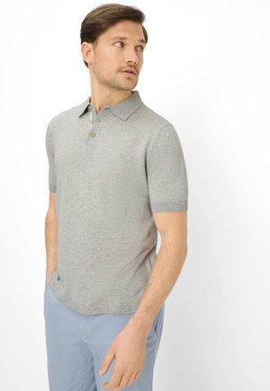 Uomo con polo a maniche corte grigio chiaro e pantaloni azzurri che guarda verso sinistra con le mani nelle tasche su sfondo bianco.