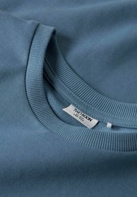 Primo piano di un maglione a collo rotondo di un leggero blu piegato, che mostra il colletto a costine e un'etichetta bianca con la taglia e il marchio all'interno del collo.