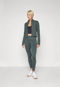 Sieviete stāv, ģērbusies zaļā plankumainā ar rāvējslēdzēju, atbilstošajās legingos, melnā īsajā topā un baltos sporta apavos uz neitrāla fona.