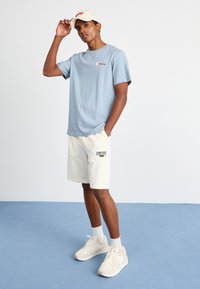 T-shirt en coton bleu clair, casquette beige avec logo, shorts crème avec un patch logo noir, baskets beiges. Le modèle se tient sur un sol bleu devant un fond uni.