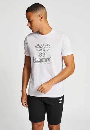 Weißes Baumwoll-T-Shirt mit einem schwarz umrandeten Logo und dem Text "hummel" auf der Brust. Kombiniert mit schwarzen Shorts, die ein kleines weißes Logo haben.