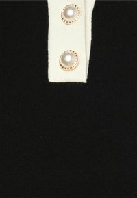 Haut en maille noire avec une patte de boutonnage crème, ornée de deux boutons ronds, semblables à des perles, entourés d'un accent doré. Texture douce, coupe ajustée.