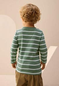 Grøn og hvid stribet langærmet skjorte med rund halsudskæring, lavet af blødt stof. Båret med khaki shorts, set fra ryggen.