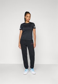 Čierne tričko s krátkym rukávom a logom na rukávoch, spárované s čiernymi teplákmi s ružovým logom a bielymi topánkami s modrými akcentmi.