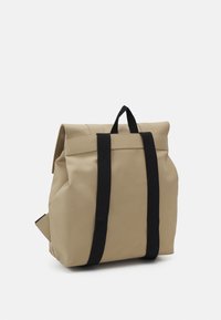 Sac à dos beige en matériau texturé, avec un système de fermeture roulée, deux sangles verticales noires et une poignée latérale pour un transport polyvalent.