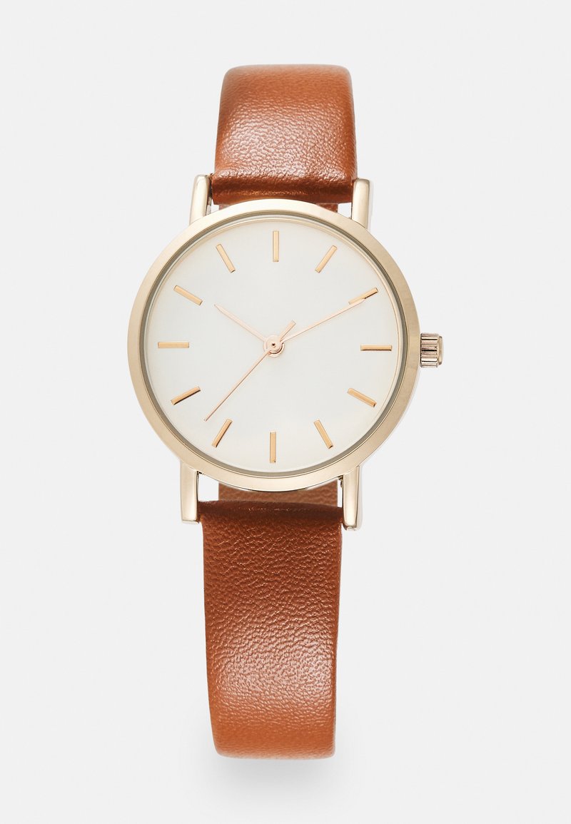 Montre avec un bracelet en cuir brun, un cadran blanc, un cadre doré et de simples marqueurs d'heure dorés. Présente une petite couronne sur le côté.
