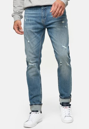 Mand iført lyseblå slidte jeans med op rullede manchetter og hvide sneakers står mod en ensfarvet baggrund.