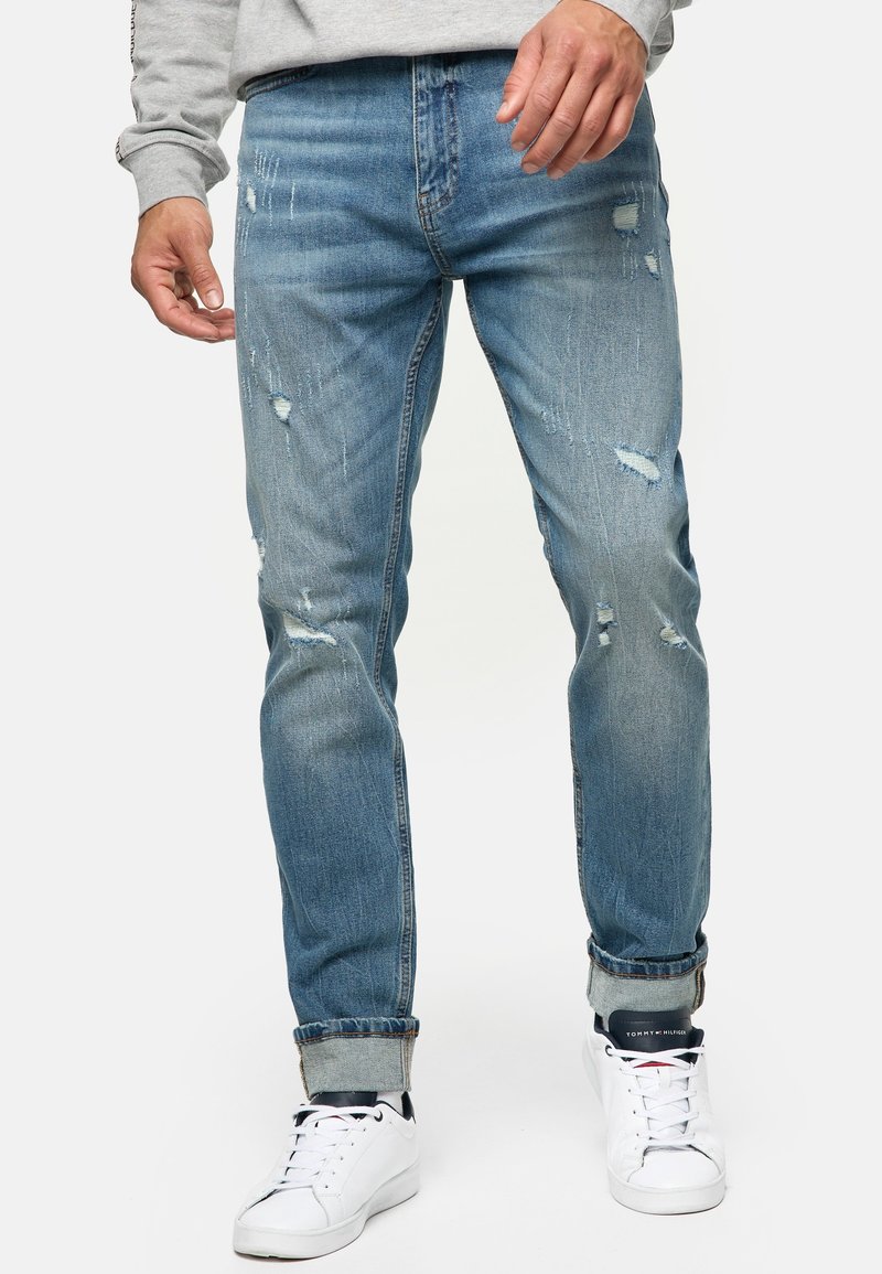 Homme portant un jean bleu clair usé avec des revers roulés et des baskets blanches, debout devant un fond uni.