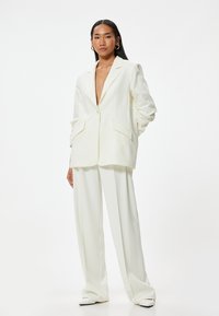 Blazer de lino blanco con un escote en V profundo y bolsillos delanteros, combinado con pantalones anchos blancos y tacones abiertos blancos.