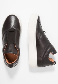 Svarta läderskor med slät yta, elastiska sidopaneler och en tjock vit gummisula. Interiören har en ljusbrun foder.