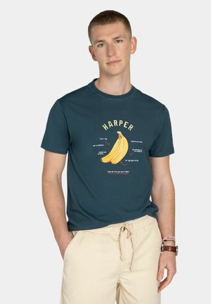 Joven con camiseta verde azulado oscura con un gráfico de un plátano amarillo y pantalones cortos beige con cordón, de pie con una mano en el bolsillo.