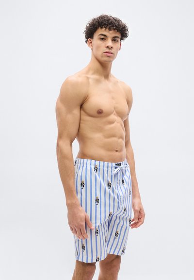 Νεαρός άνδρας με σγουρά μαλλιά, φορώντας μπλε και λευκά ριγέ σορτς με σχέδια αρκουδάκια, στέκεται μπροστά σε ένα απλό φωτεινό φόντο.