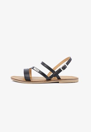 Sandal in pelle nera con due cinturini incrociati, cinturino alla caviglia regolabile con fibbia e suola piatta beige. Dettaglio del marchio sul cinturino.