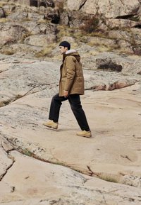 Jachetă brună izolată cu glugă, hanorac gri dedesubt, pantaloni negri și cizme bej, pe un fundal de teren stâncos.