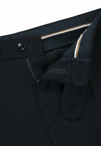 Svarta texturerade byxor i tyg med dragkedja, två svarta knappar och en randig midjebanddetalj i guld och vitt.