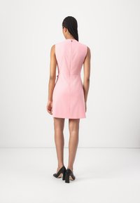 Robe courte rose au design ajusté, sans manches, avec une fermeture éclair dans le dos. Le modèle porte des chaussures à talons bloc noires. Style minimaliste.