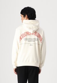 Haut à capuche crème avec un grand texte graphique rouge au dos. Le design comprend une poche kangourou et des poignets et un ourlet côtelés.