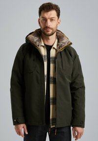 Donkergroene jacket met een met bont gevoerde kraag, borstzakken en een zwarte binnenkant, gelaagd over een romig geblokt shirt. Voorzien van rits- en drukknoopsluitingen.