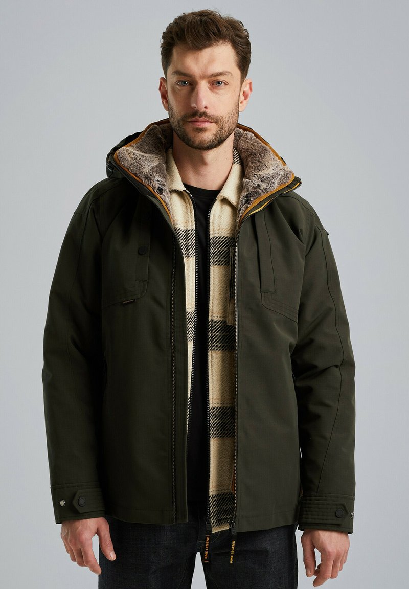 Donkergroene jacket met een met bont gevoerde kraag, borstzakken en een zwarte binnenkant, gelaagd over een romig geblokt shirt. Voorzien van rits- en drukknoopsluitingen.