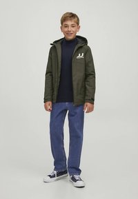 Πράσινο μπουφάν με κουκούλα και φερμουάρ στο μπροστινό μέρος, με λεπτομέρεια λογότυπου, συνδυασμένο με μπλε ζιβάγκο, μπλε τζιν και μπλε αθλητικά παπούτσια με λευκές σόλες.